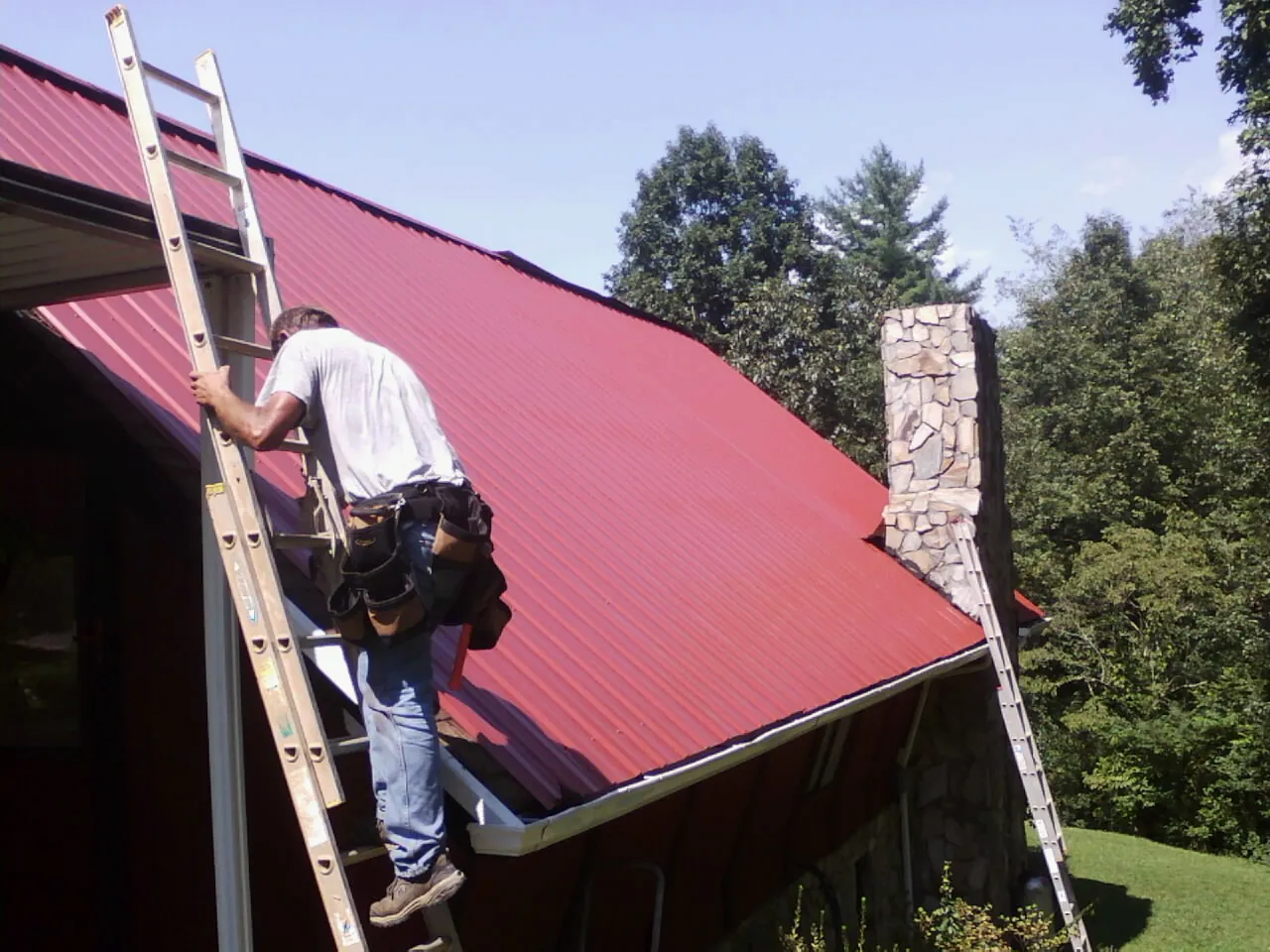 Roofer on a ladder beside a red metal roof and stone chimney — Soffit & Fascia Repair by Roofing Brothers in Thompson Ridge